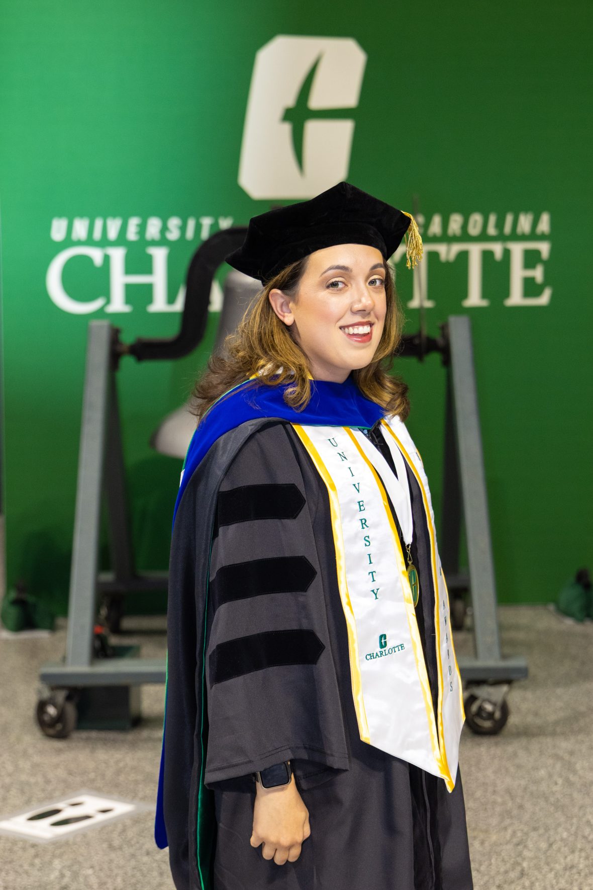 Kaitlin Klotz posing in front of the victory bell in her doctoral regalia and graduation stoles.