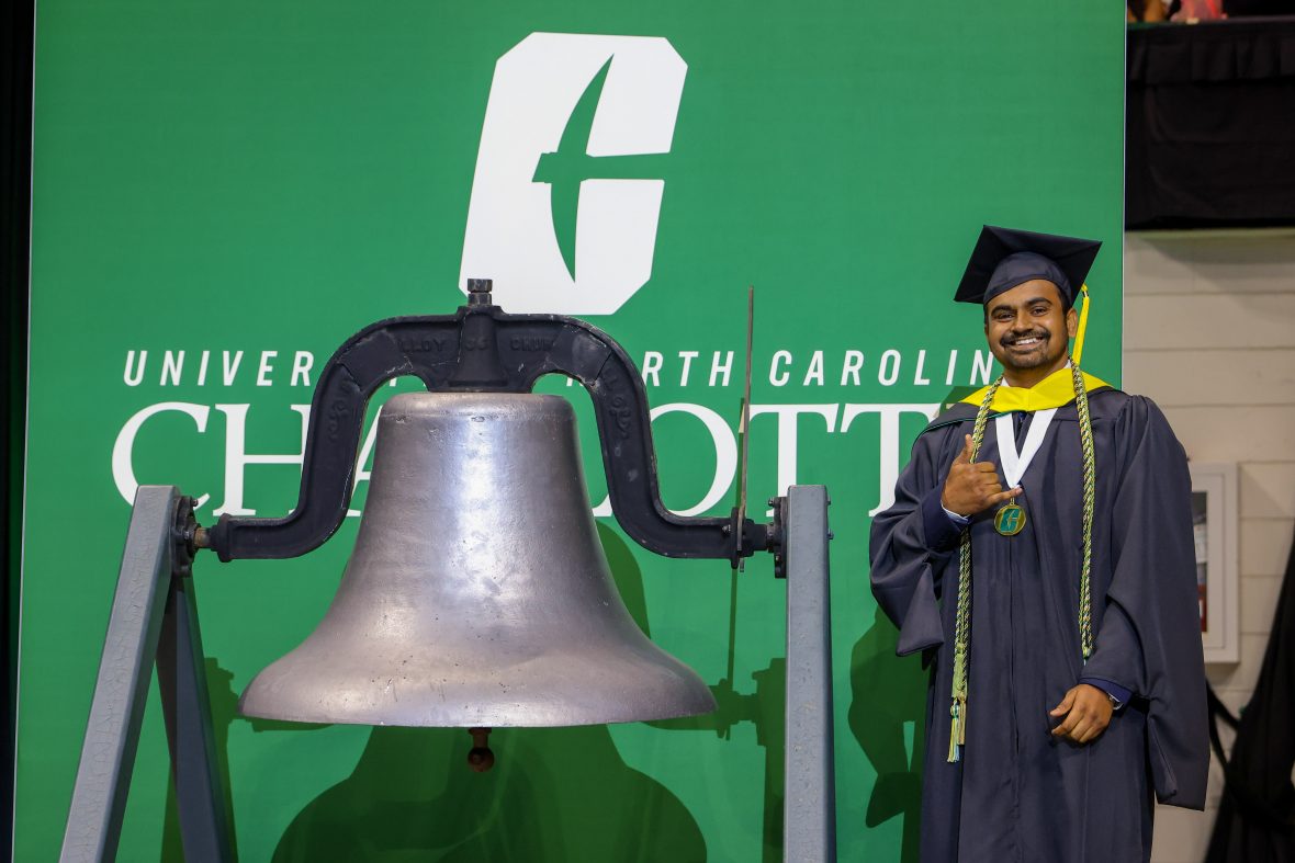 Shailesh Kumar posing next to the victory bell with the Niner "Picks Up" hand gesture, wearing his Master's cap and gown.